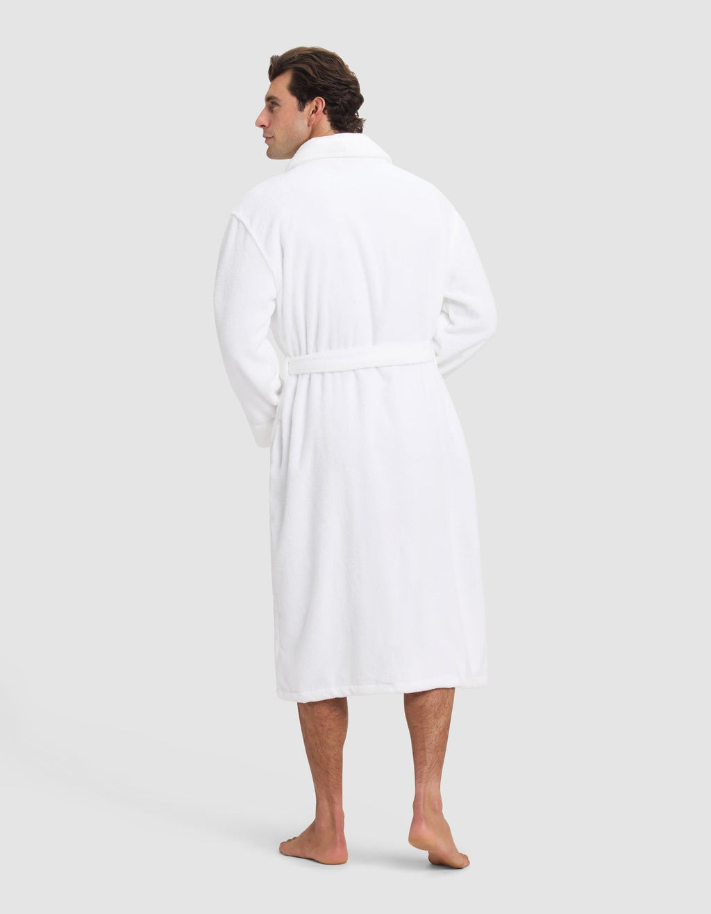 A man stands barefoot facing away, wearing the Luxe Bath Robe by Cozy Earth, featuring a belt, against a plain light gray background.|Color:White