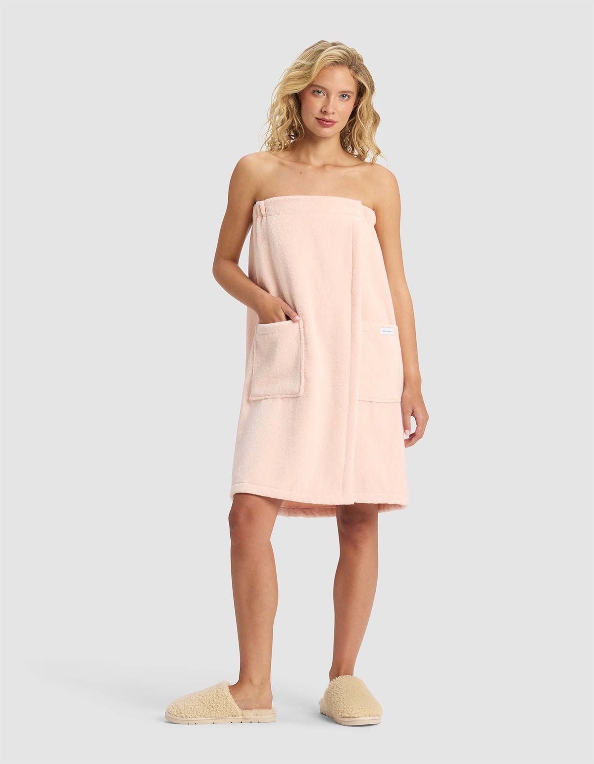 A woman with blonde hair wears the Cozy Earth Luxe Bath Wrap in light pink, featuring two front pockets, along with beige slippers, standing against a plain light gray background.|Color:Blossom