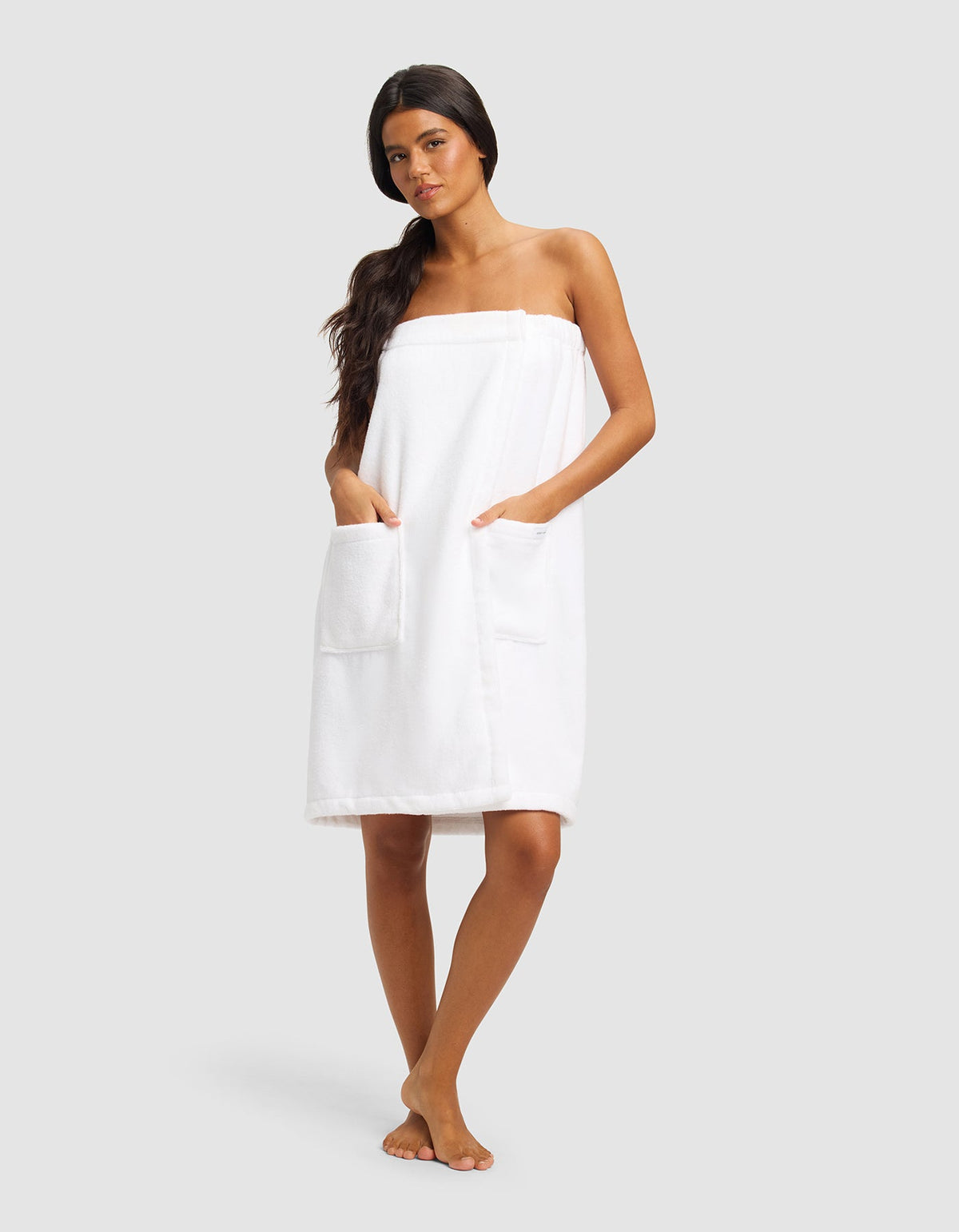 A woman with long dark hair stands barefoot against a plain background, wearing Cozy Earth's Luxe Bath Wrap featuring two large front pockets.|Color:White