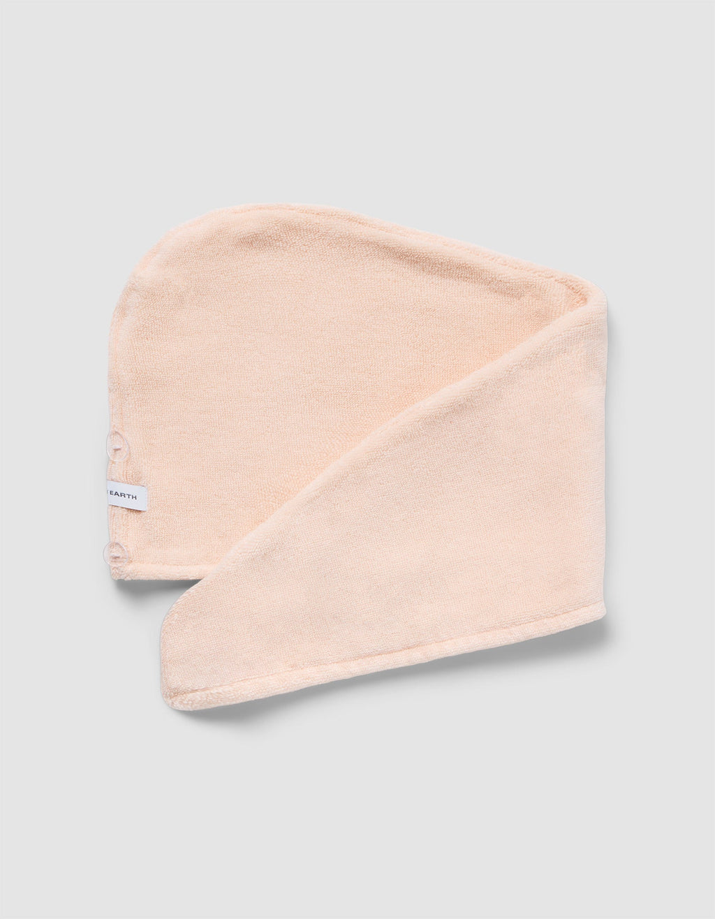The Luxe Hair Towel by Cozy Earth is a soft, light beige microfiber towel with a white label and two snap buttons. Displayed partially folded on a light gray background, it highlights its rectangular shape and smooth texture.|Color:Blossom