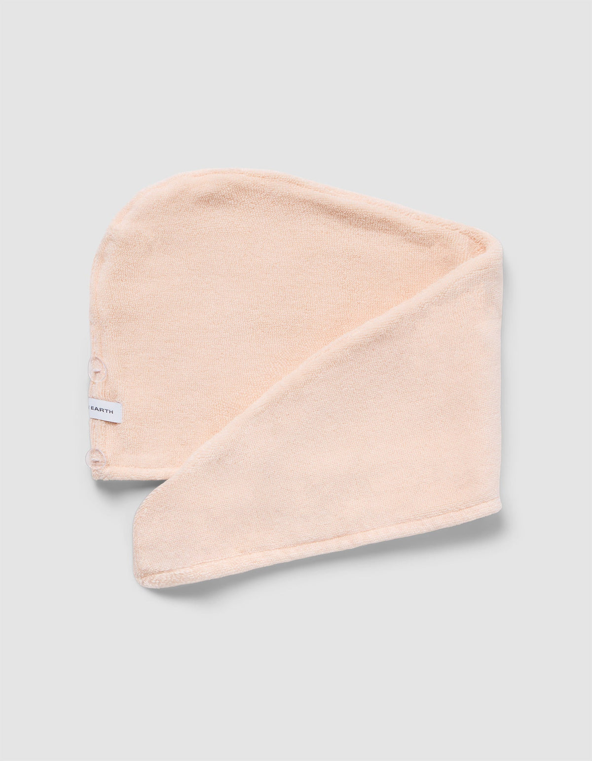 The Luxe Hair Towel by Cozy Earth is a soft, light beige microfiber towel with a white label and two snap buttons. Displayed partially folded on a light gray background, it highlights its rectangular shape and smooth texture.|Color:Blossom