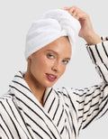 A woman smiles softly, touching the Cozy Earth Luxe Hair Towel wrapped around her head, while wearing a white bathrobe with black stripes against a plain light background. |Color:White
