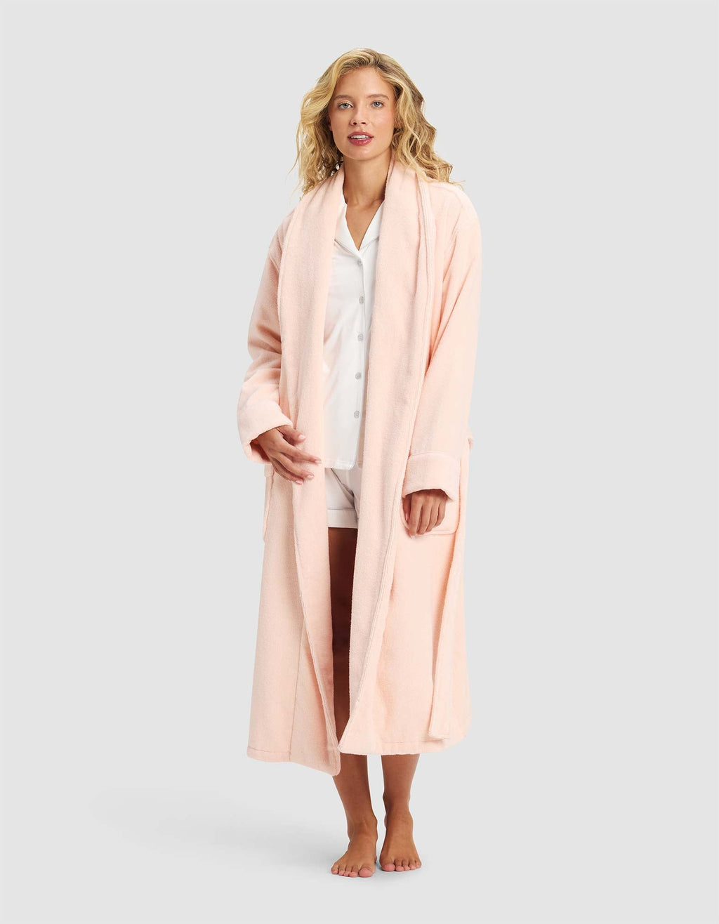 A woman stands barefoot against a plain light gray background, wearing Cozy Earth's Luxe Bath Robe in light pink over white pajama shorts and a matching button-up shirt.|Color:Blossom