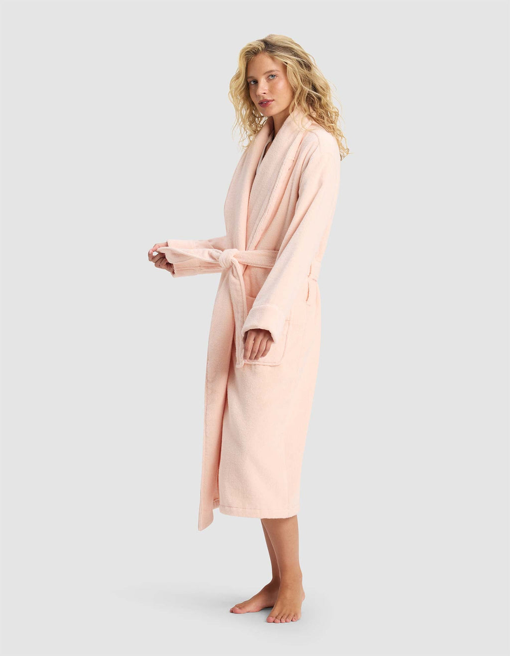 A woman with curly blonde hair wears the Cozy Earth Luxe Bath Robe in light pink, tied at the waist with two front pockets, standing barefoot against a plain light gray background.|Color:Blossom