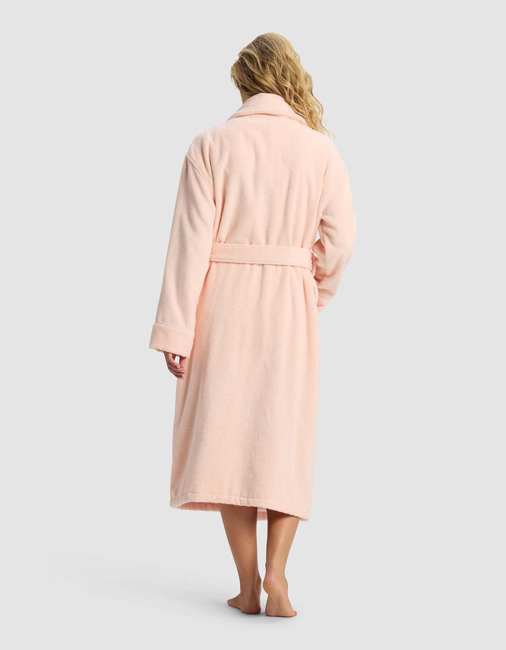 A person with long, wavy blonde hair stands barefoot, facing away, wearing the Cozy Earth Luxe Bath Robe in light pink with a belt against a plain light gray background.|Color:Blossom