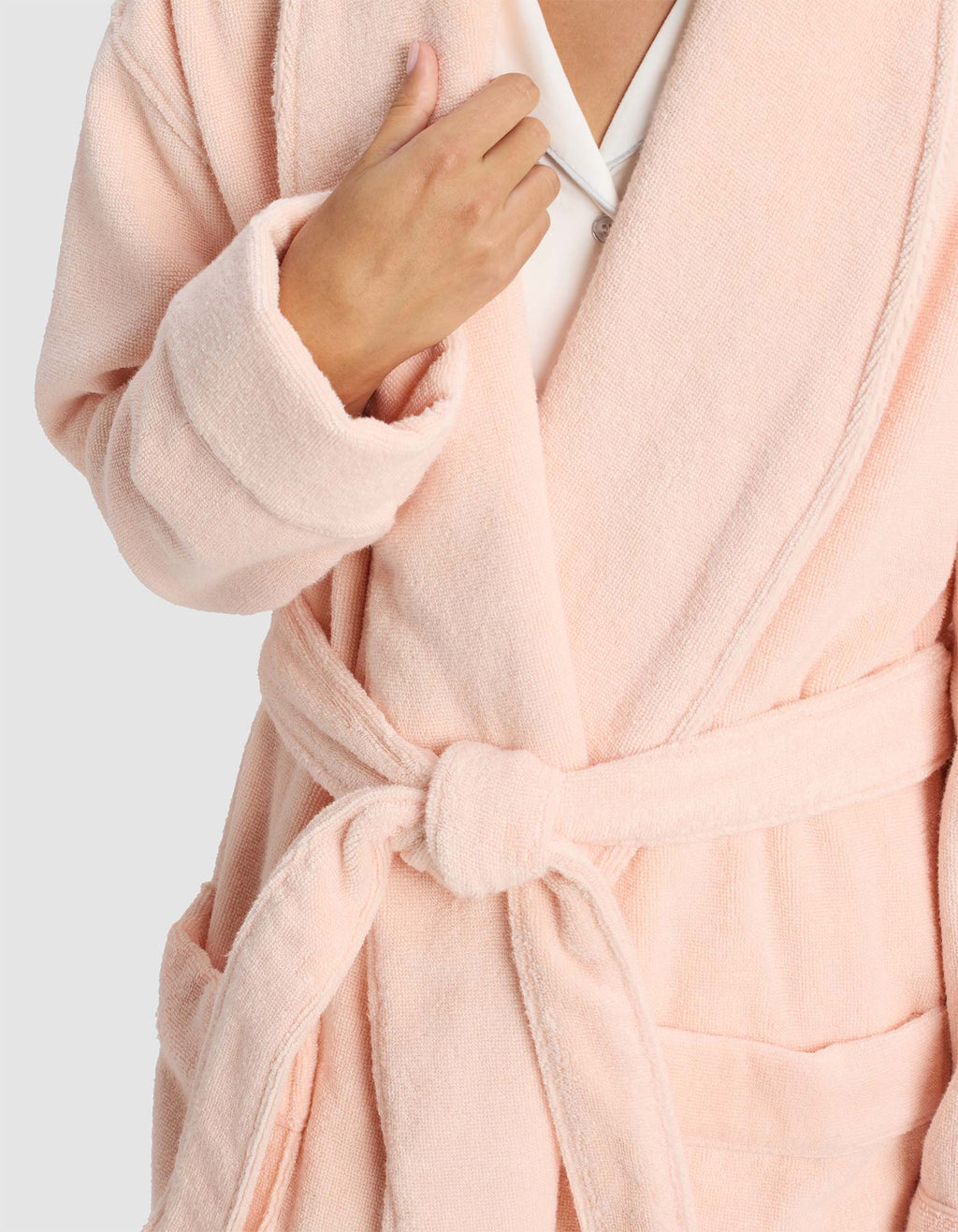 A person wears the Luxe Bath Robe by Cozy Earth in light pink, tied at the waist and holding the lapel. The background is plain and light-colored, with their face partially out of frame.|Color:Blossom