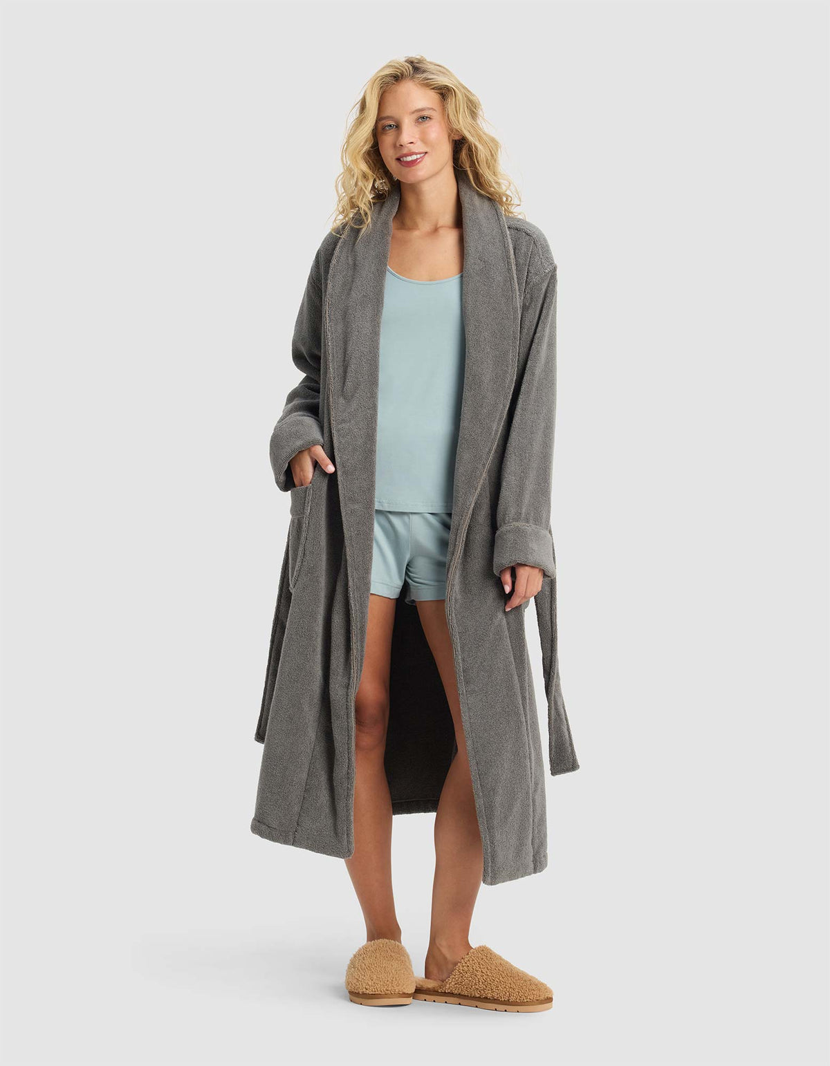 A woman smiles with her hands in the pockets of a Luxe Bath Robe by Cozy Earth, layering it over light blue pajamas and tan slippers against a plain light gray background.|Color:Charcoal