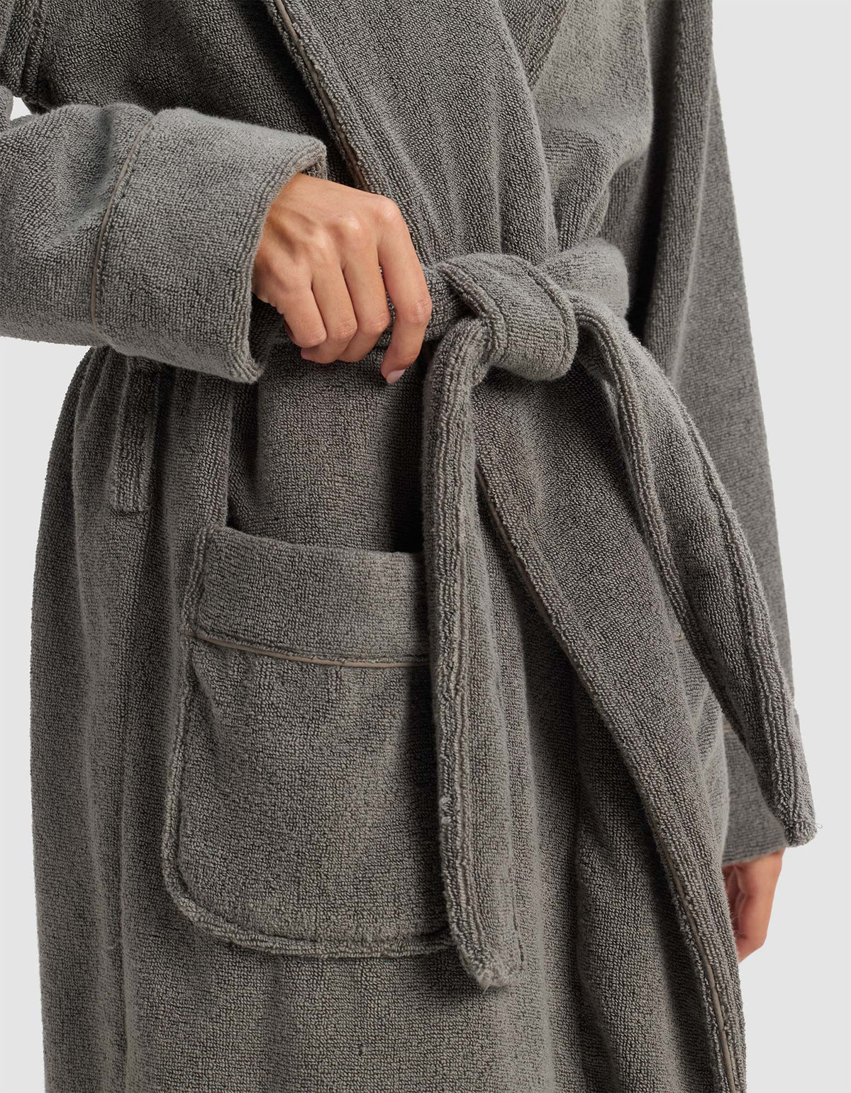 A person in the Cozy Earth Luxe Bath Robe ties the waist belt; one hand loops the belt, and the plush robe features a spacious front pocket.|Color:Charcoal