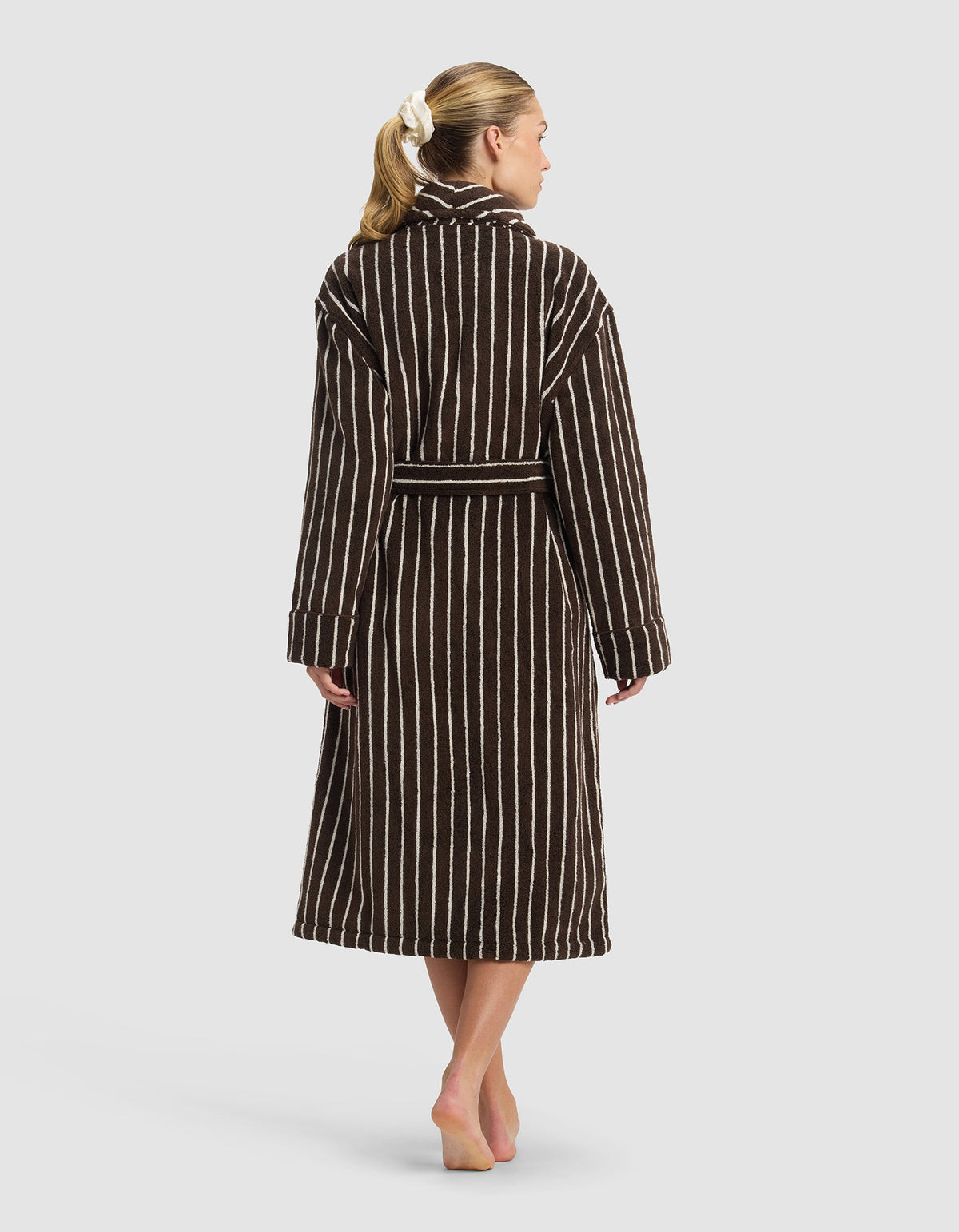 A person with light skin and blonde hair in a white scrunchie stands barefoot, back to camera, wearing the Cozy Earth Striped Luxe Bath Robe—a long, dark brown robe with vertical white stripes—against a plain light background.|Color:Espresso Coconut Stripe