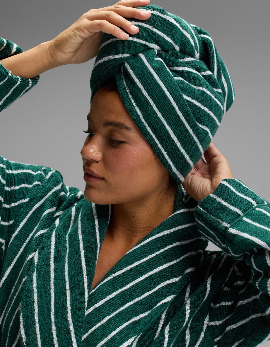 A woman wearing a green and white striped robe adjusts the Luxe Hair Towel by Cozy Earth on her head, looking to the side against a gray background.|Color:Uniform with Seashell Stripe