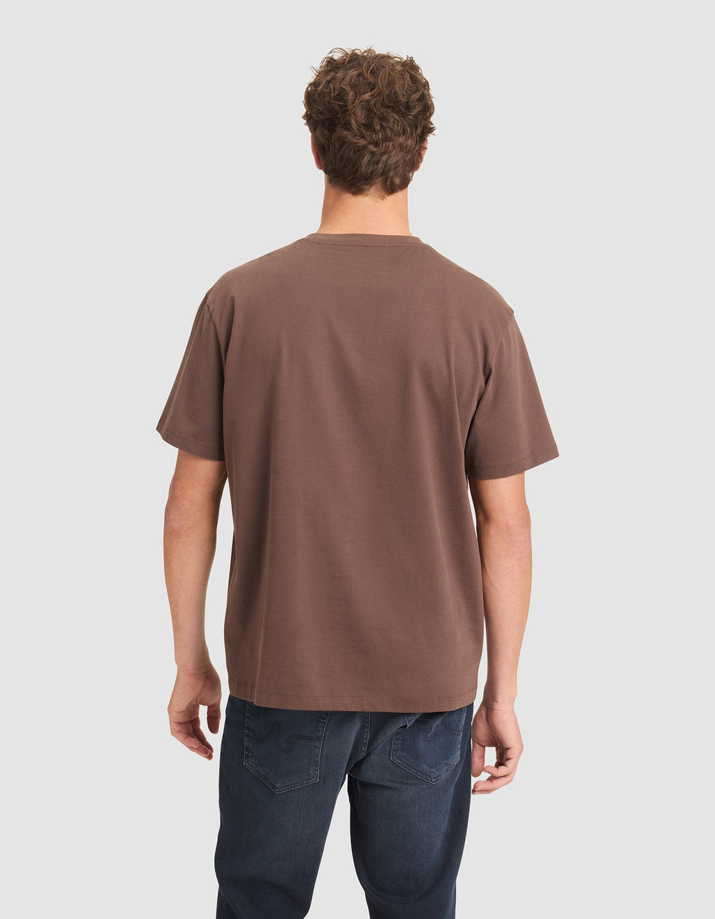 A man with short curly hair, seen from behind, wears a Cozy Earth Men's All Day Tee in brown and dark blue jeans against a plain light background. |Color:Cacao