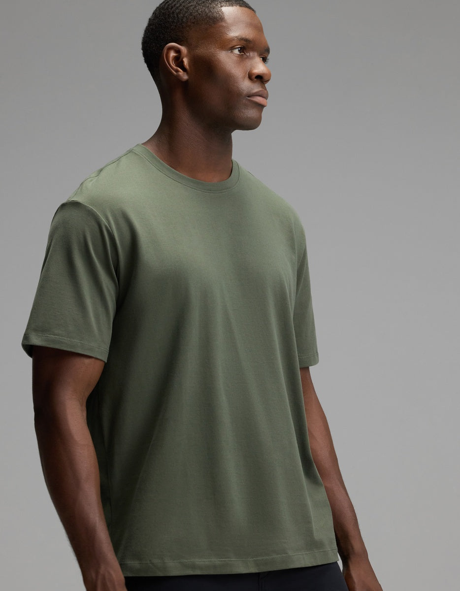 A man wearing the Cozy Earth Men’s All Day Tee in olive green stands against a neutral gray background, looking off to the side with a serious expression.|Color:Olive