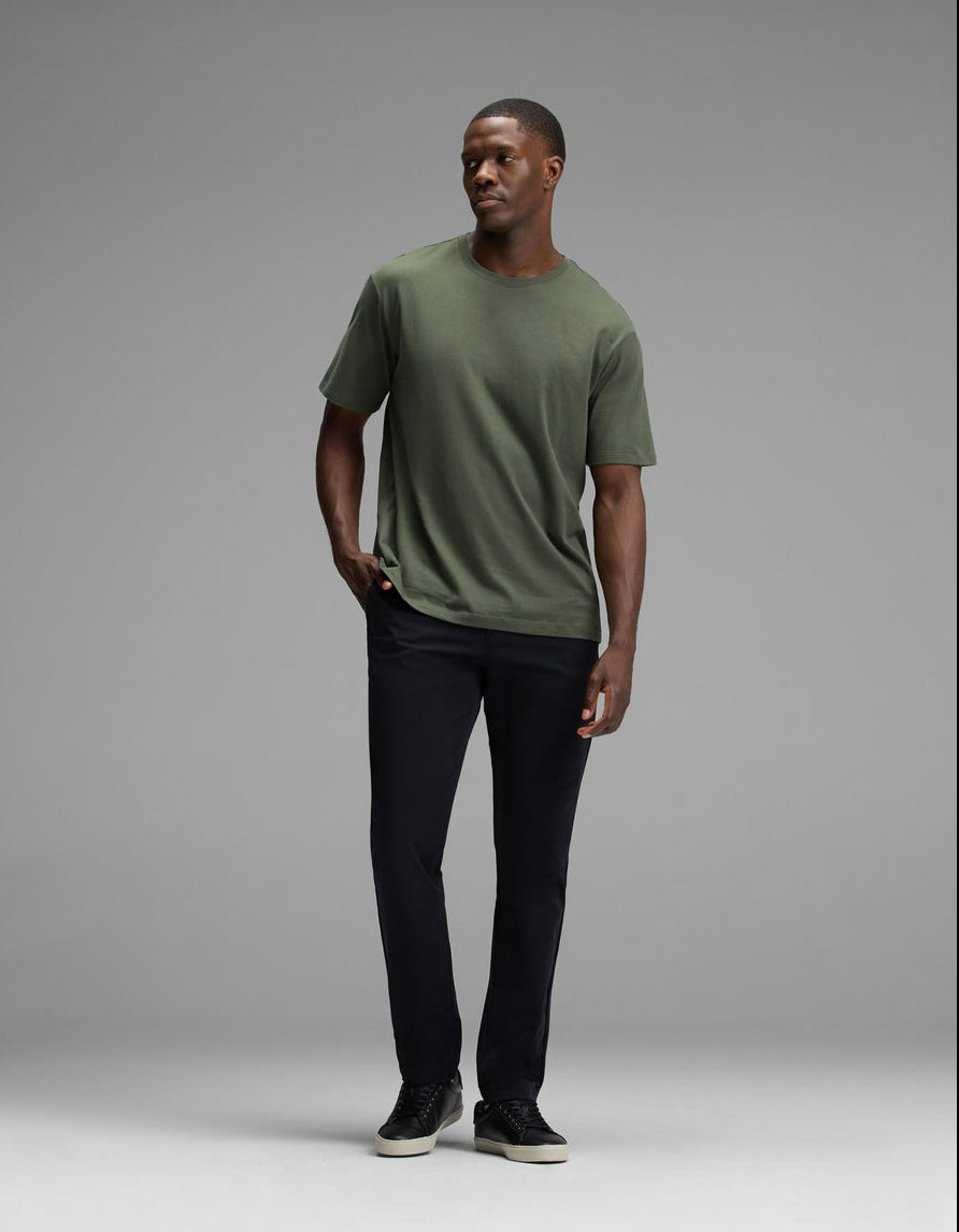 A man stands against a gray background, wearing the Cozy Earth Men’s All Day Tee in green, paired with black pants and black sneakers. He has one hand in his pocket and looks off to the side.|Color:Olive