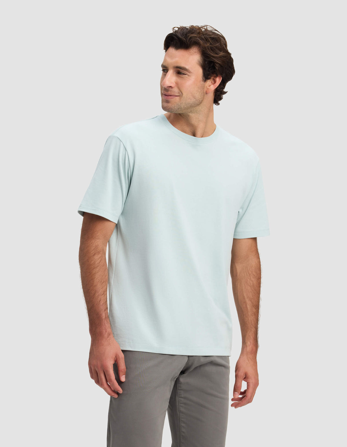 A man with dark hair models the Cozy Earth Men's All Day Tee in light blue and gray pants, standing against a plain light gray background, looking slightly to his left and smiling.|Color:Tide