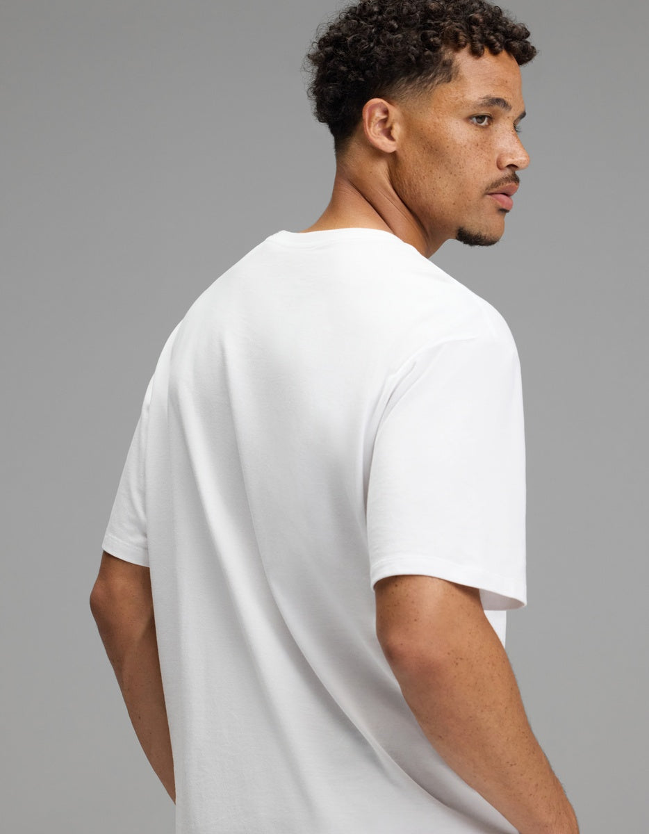 A man with short curly hair and a trimmed beard, wearing the Cozy Earth Men’s All Day Tee in plain white, turns to look over his right shoulder against a neutral gray background.|Color:White
