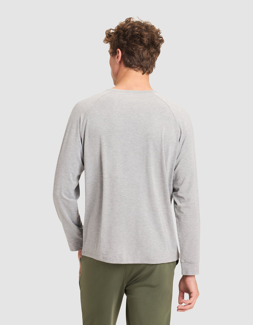 A man with curly hair stands with his back to the camera, wearing a Cozy Earth Men's Bamboo Stretch-Knit Long Sleeve in light gray and olive green pants against a plain light background. |Color:Heather Grey