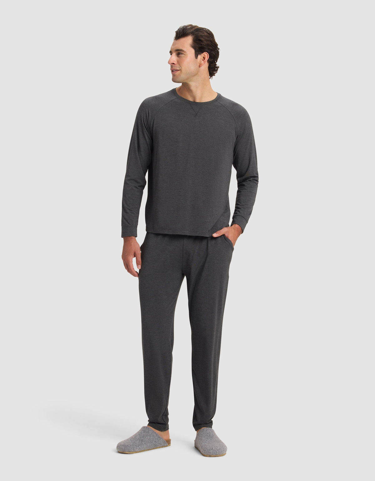A man with short dark hair smiles in a plain white setting, wearing Cozy Earth Men's Bamboo Stretch-Knit Pajama Pants and matching dark gray long-sleeve top, paired with gray slippers and one hand in his pocket.|Color:Charcoal