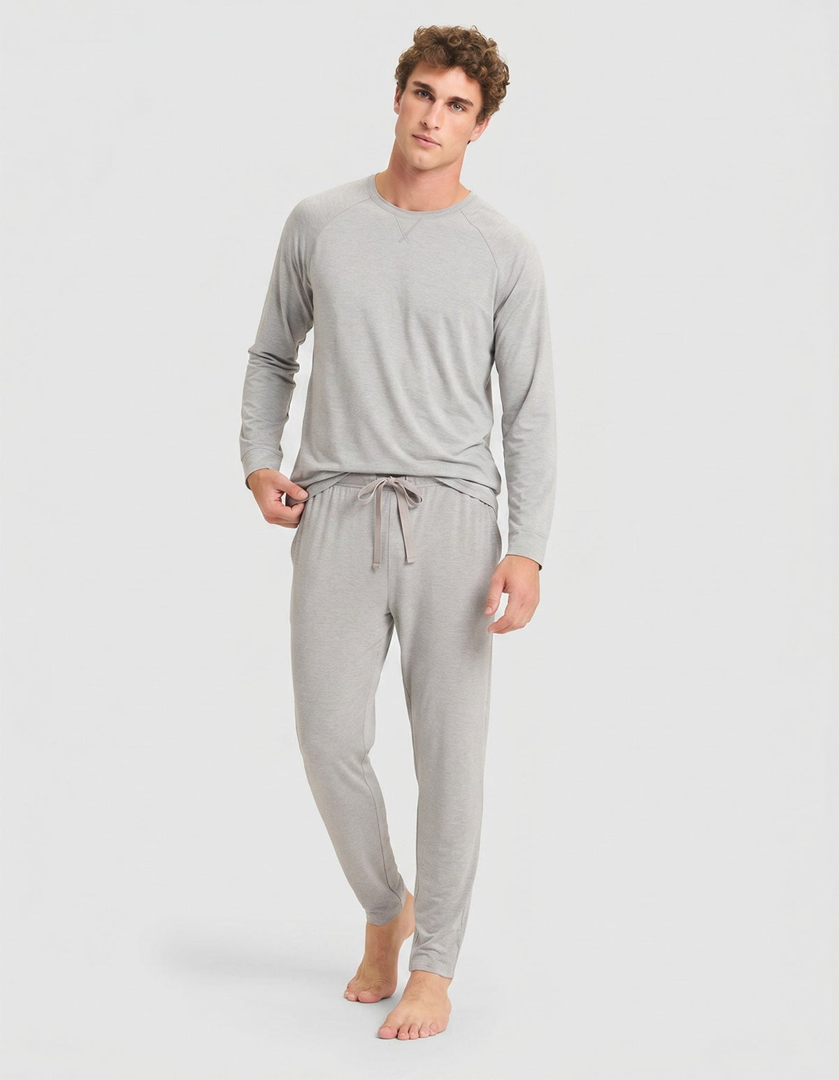 A person stands barefoot against a plain light background, wearing the Cozy Earth Men's Bamboo Stretch-Knit Long Sleeve Pajama Set in Heather Grey.|Color:Heather Grey