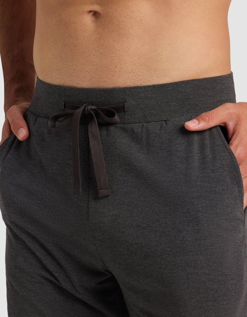A close-up of a person wearing Cozy Earth's Men's Bamboo Stretch-Knit Pajama Short in dark gray, with hands in the pockets. The shirtless model is shown from waist to upper thighs, highlighting the comfortable stretch-knit fabric.|Color:Charcoal