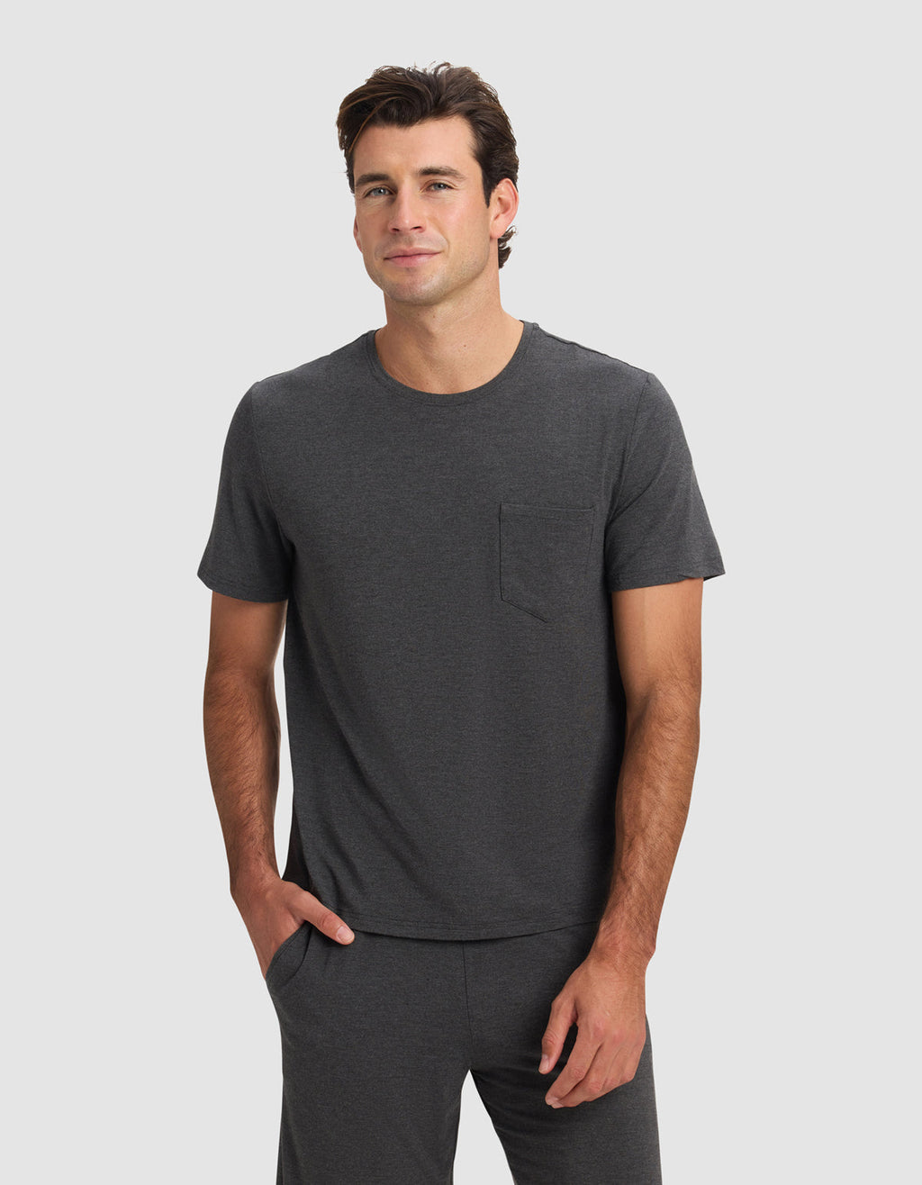 A man wearing the Cozy Earth Men's Bamboo Stretch-Knit Lounge Tee in charcoal gray, featuring short sleeves and a chest pocket, stands against a light gray background with one hand in his pocket, smiling slightly.|Color:Charcoal