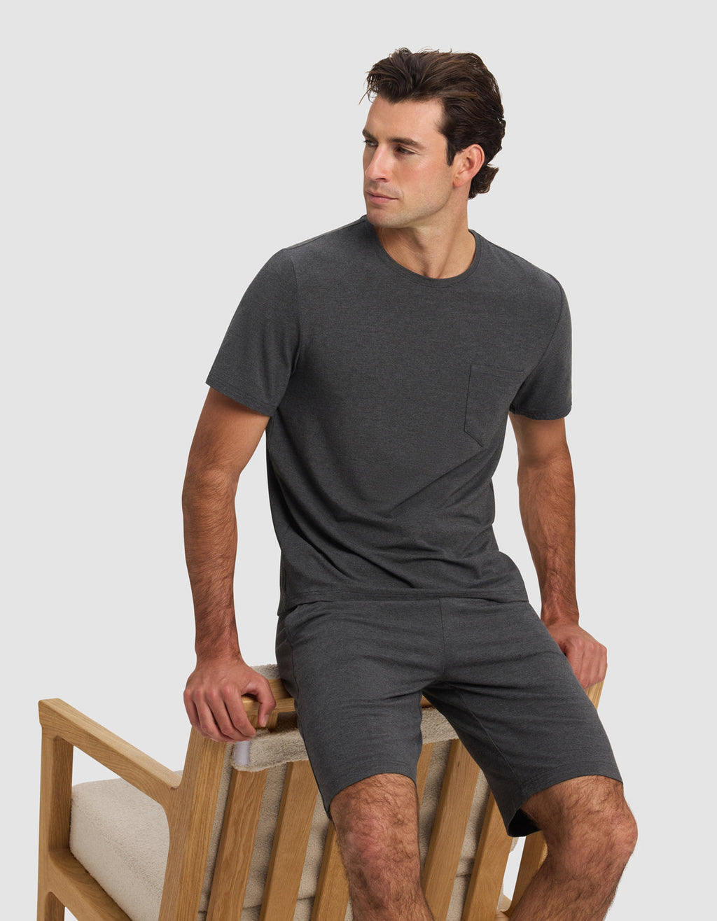 A man wears the Cozy Earth Men's Bamboo Stretch-Knit Lounge Tee in dark gray, paired with matching shorts, while sitting sideways on a light wooden chair with beige cushions against a plain light background.|Color:Charcoal