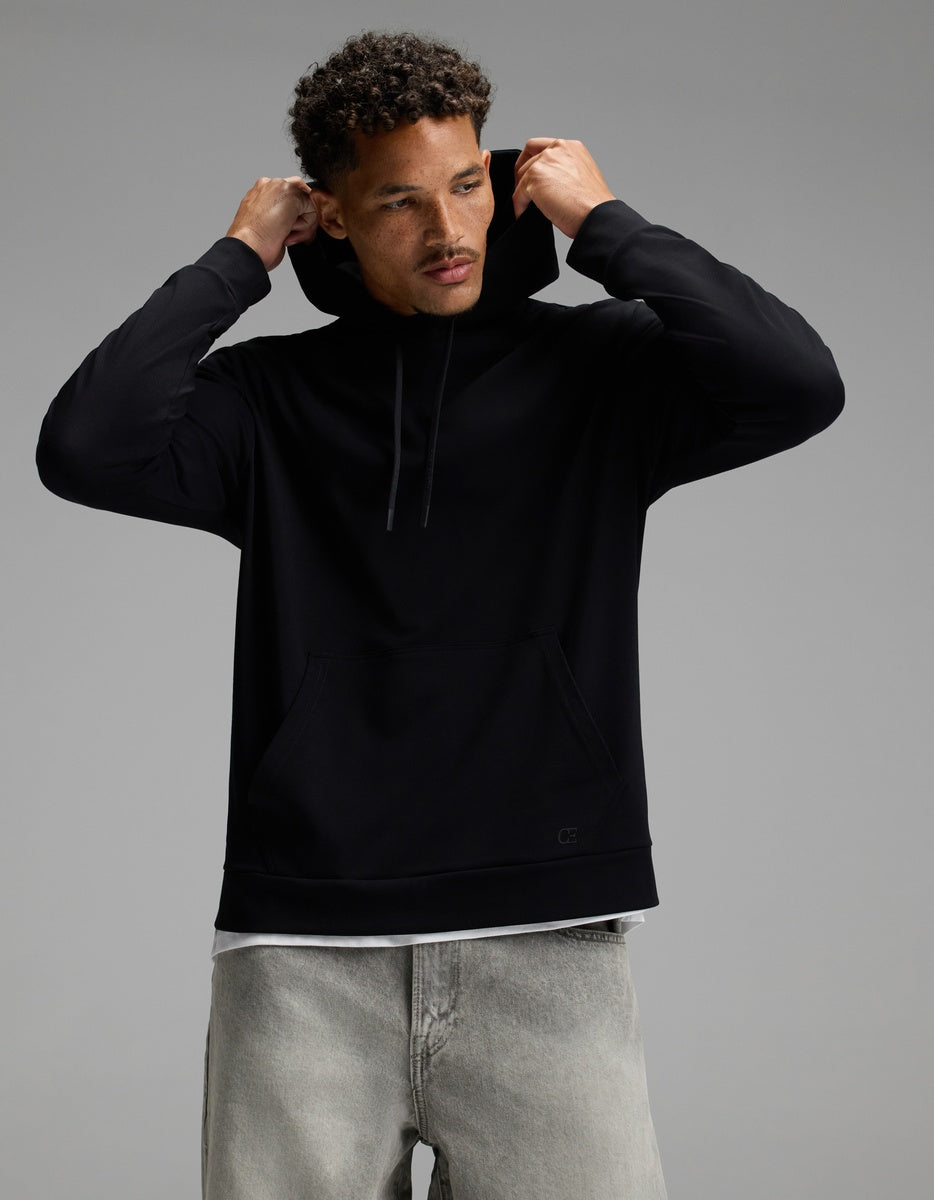 A man wearing the Cozy Earth Men’s Brushed Bamboo Hoodie in black and light gray jeans adjusts the hood while looking slightly down against a plain gray background.|Color:Black