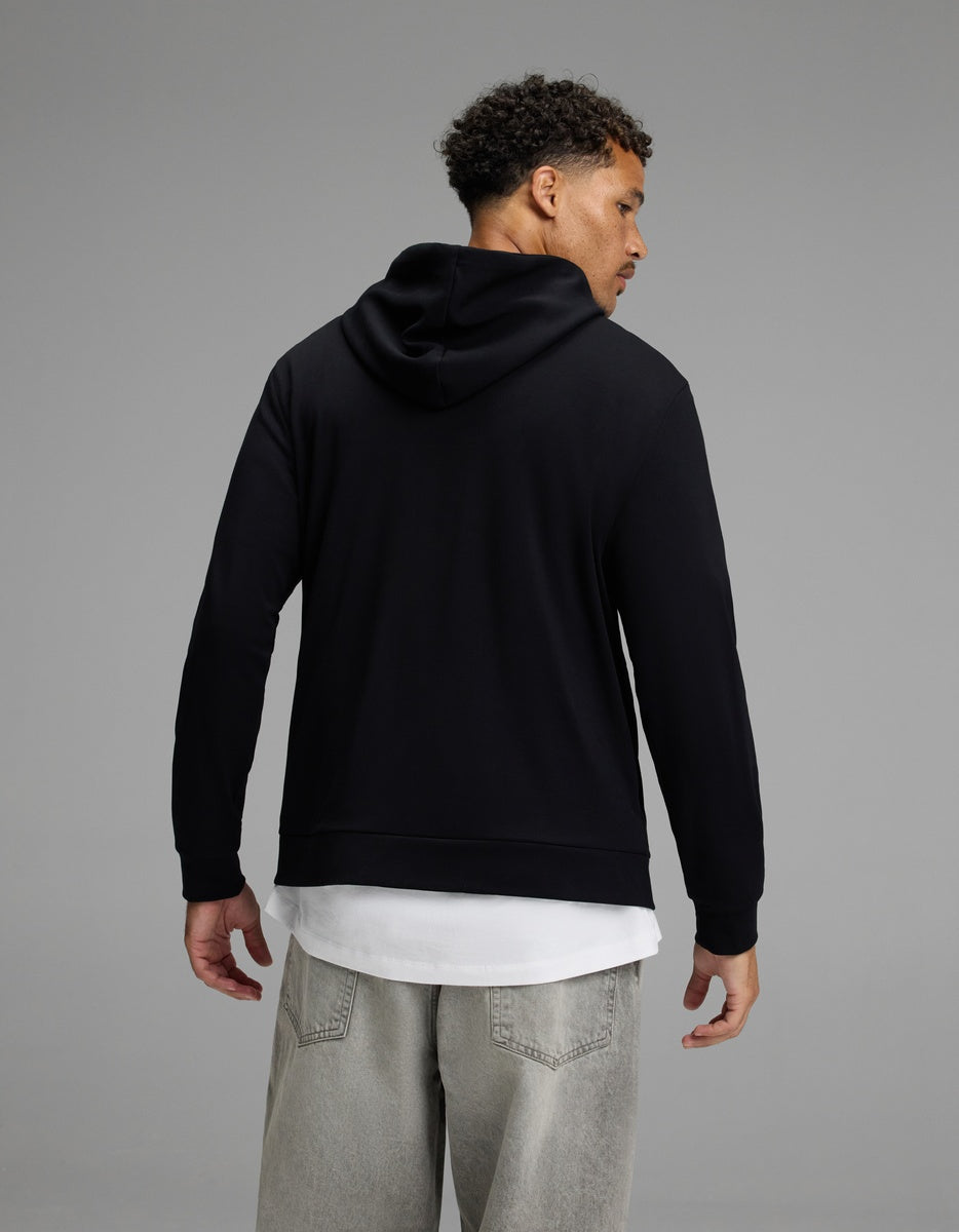 A man stands facing away, wearing the Cozy Earth Men’s Brushed Bamboo Hoodie in black over a white shirt and light gray jeans, set against a plain gray background.|Color:Black