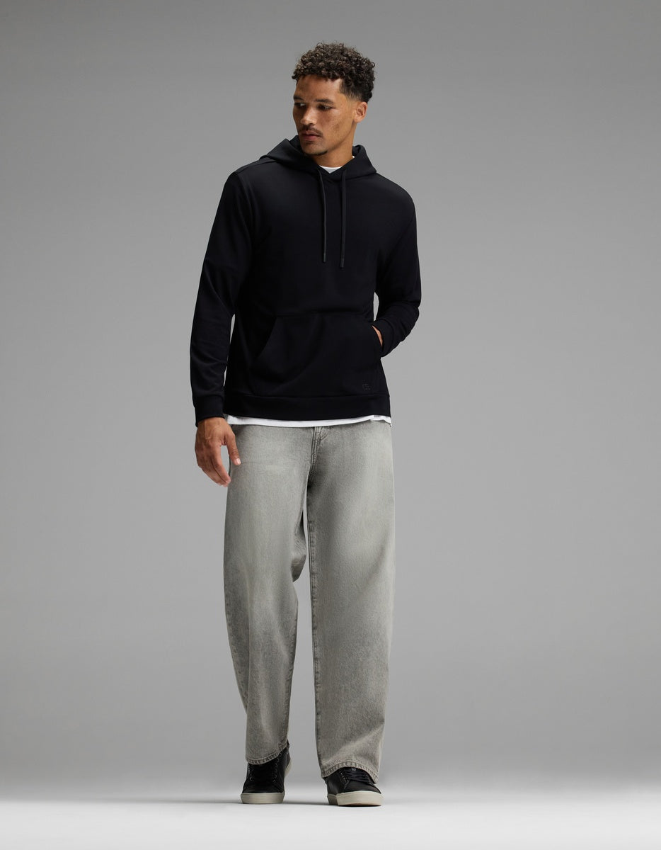 A man stands against a plain gray background, looking to his left with one hand in the pocket of his Cozy Earth Men’s Brushed Bamboo Hoodie, layered over a white shirt and paired with loose-fitting gray jeans.|Color:Black