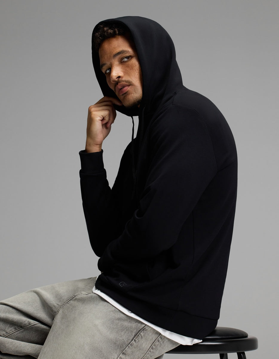A man wearing the Cozy Earth Men’s Brushed Bamboo Hoodie and gray jeans sits on a stool against a plain gray background, looking seriously at the camera with one hand near his chin.|Color:Black