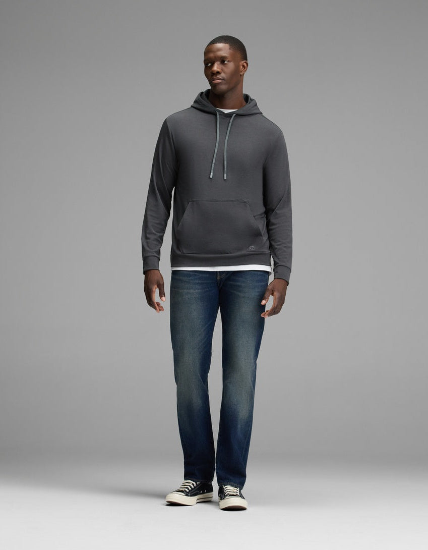 A man stands against a plain gray background wearing the Cozy Earth Men’s Brushed Bamboo Hoodie in dark gray, paired with blue jeans and black-and-white sneakers. He faces slightly to the side with his hands relaxed by his sides.|Color:Charcoal