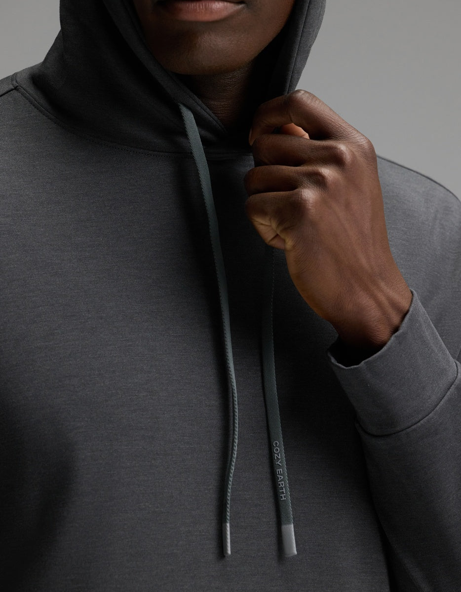 A person wearing the Cozy Earth Men’s Brushed Bamboo Hoodie in dark gray holds the hood’s drawstring—featuring a subtle logo—near their chin, with a plain gray background.|Color:Charcoal