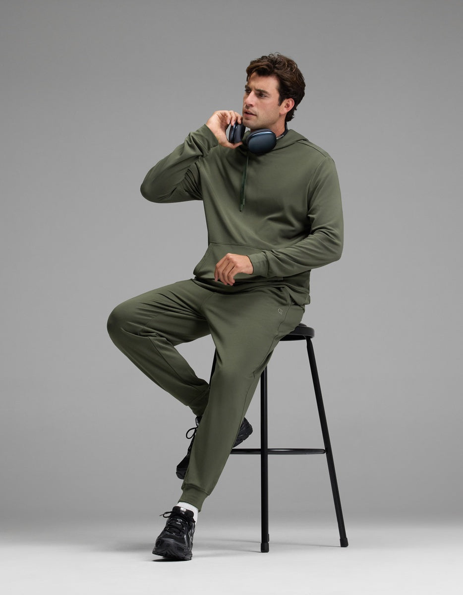 A man wearing the Cozy Earth Men’s Brushed Bamboo Hoodie & Jogger Set in olive green sits on a black stool, holding a phone to his ear with headphones resting around his neck, against a plain gray background.|Color:Olive
