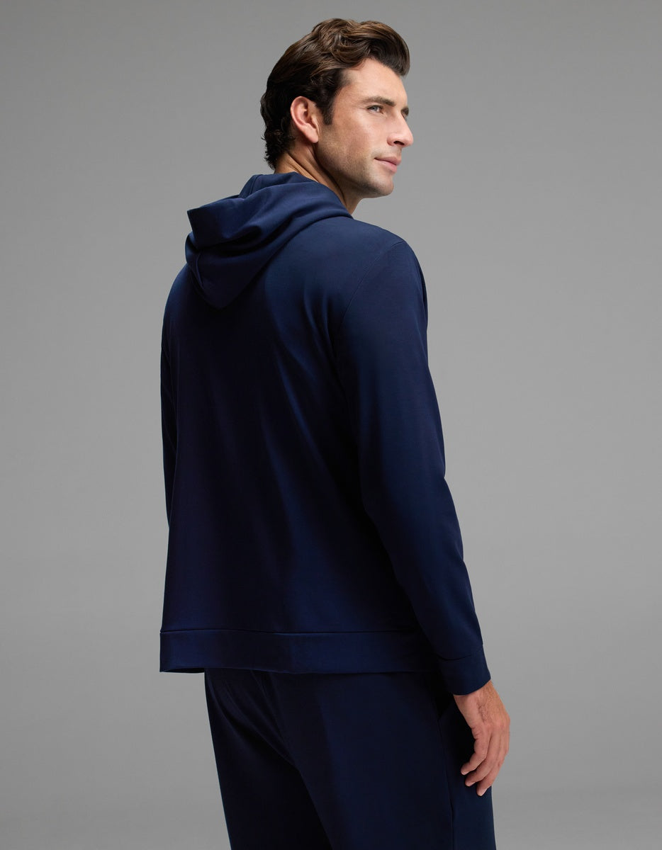 A man with brown hair wears the Cozy Earth Men’s Brushed Bamboo Hoodie in navy blue, paired with matching pants, as he stands facing away and looks over his shoulder against a plain gray background.|Color:Navy