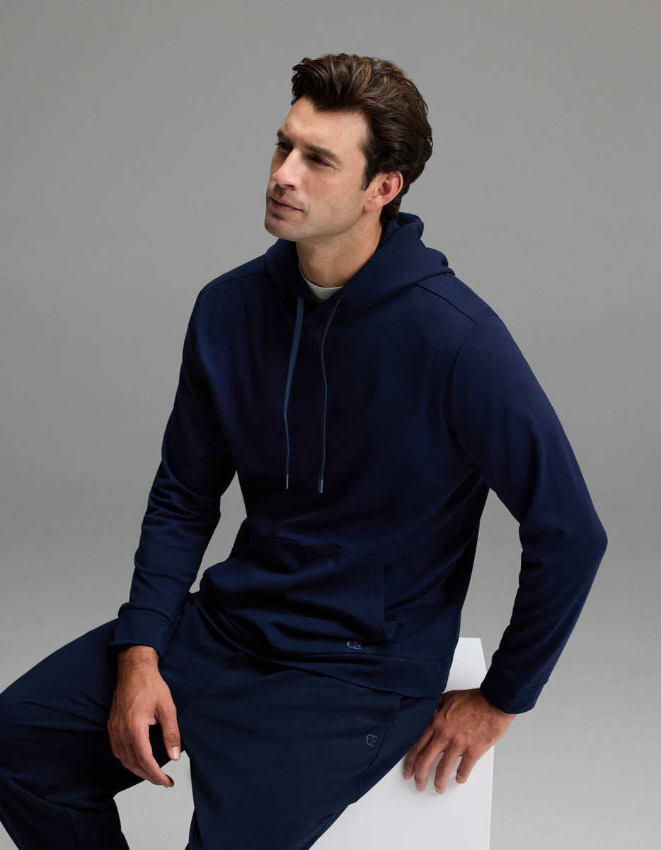 A man with short brown hair, dressed in the Cozy Earth Men’s Brushed Bamboo Hoodie and matching pants in navy blue, sits on a white block against a plain gray background.|Color:Navy