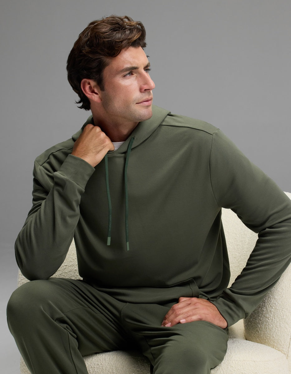 A man in the Cozy Earth Men’s Brushed Bamboo Hoodie and matching pants sits on a light-colored chair, gazing thoughtfully to his left with one hand touching his neck and the other resting on his knee.|Color:Olive