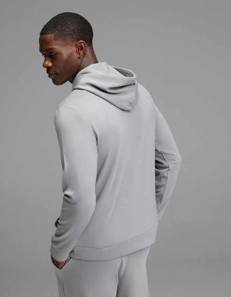 A person wearing the Cozy Earth Men’s Brushed Bamboo Hoodie in light gray, paired with matching pants, stands with hands in pockets and faces slightly away against a plain gray background.|Color:Stone