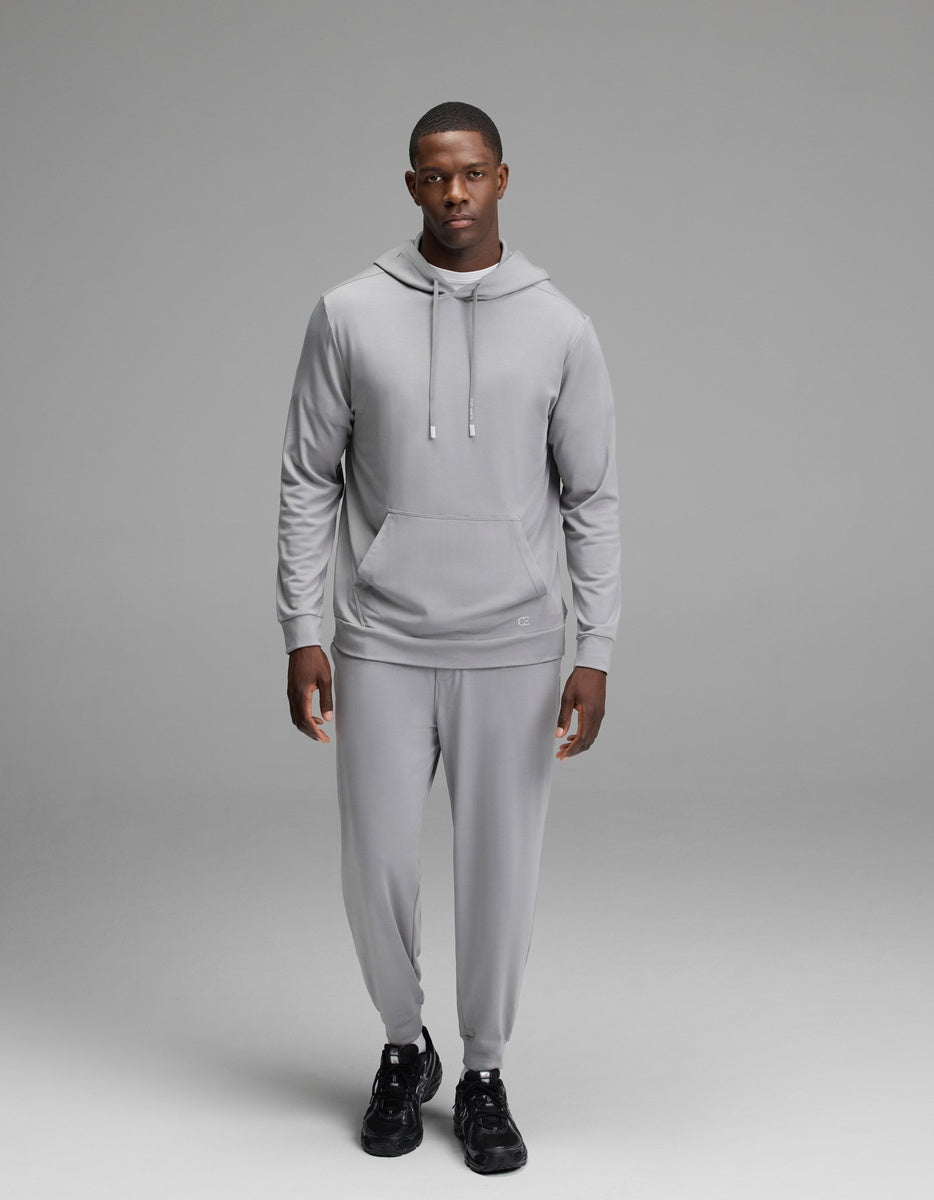 A man wearing the Cozy Earth Men’s Brushed Bamboo Hoodie in light gray and matching pants stands against a plain gray background, looking forward with a neutral expression. He is also wearing black sneakers.|Color:Stone