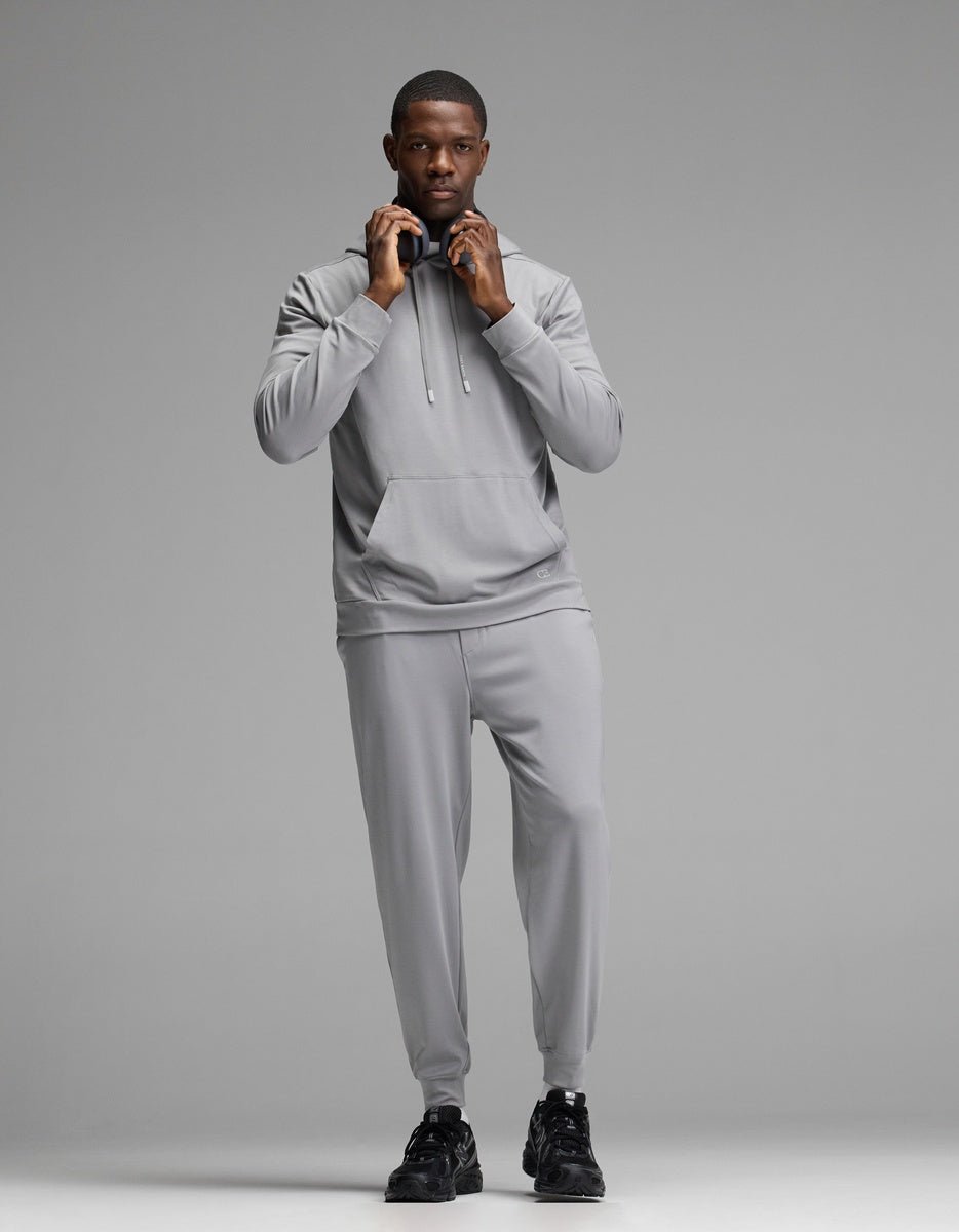 A person stands facing forward against a plain gray background, wearing Cozy Earth’s Men’s Brushed Bamboo Hoodie in light gray along with matching joggers and black sneakers, adjusting the hood drawstrings.|Color:Stone