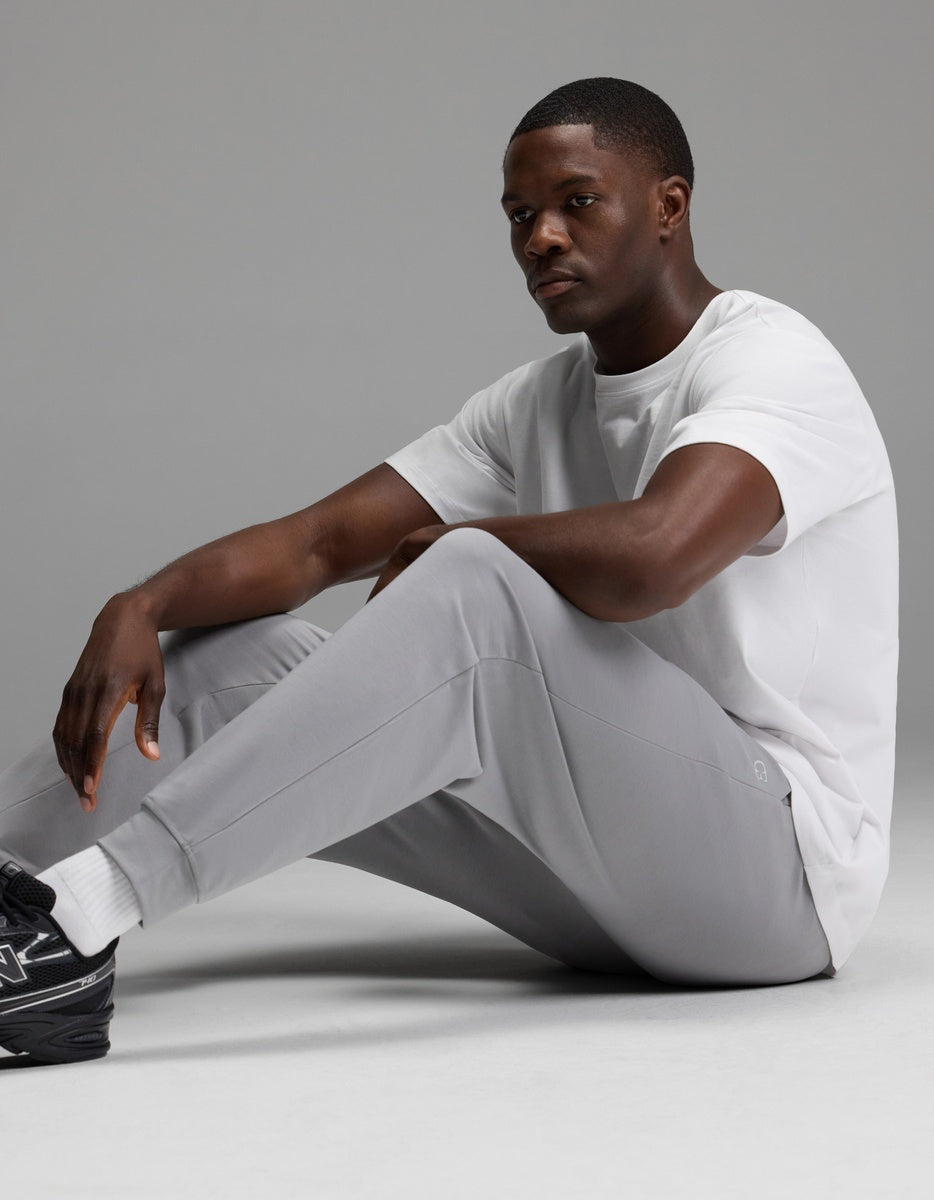 A man wearing Cozy Earth’s Men’s Brushed Bamboo Joggers in light gray, paired with a white t-shirt, white socks, and black sneakers, sits on the floor against a plain gray background.|Color:Stone