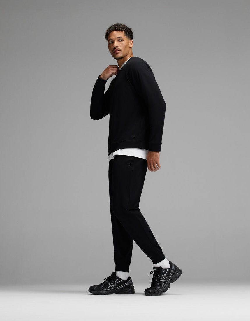 A man stands in a studio wearing the Cozy Earth Men’s Brushed Bamboo Jogger Set in black, paired with black sneakers and a white undershirt. He looks to the side with one hand near his neck, against a plain gray background.|Color:Black