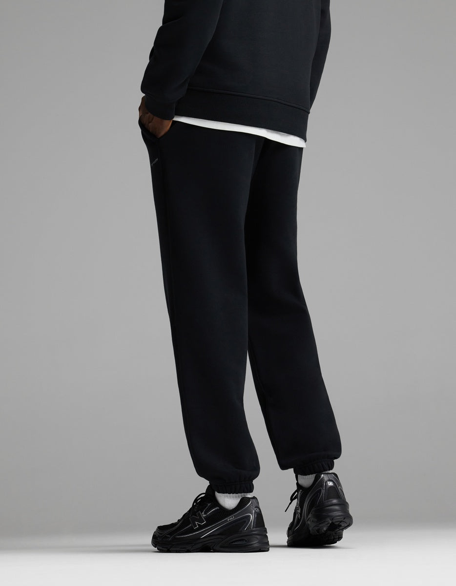 A person wearing Cozy Earth Men’s Cityscape Sweatpant in black stands facing away, with only their legs and lower torso visible, against a plain gray background.|Color:Jet Black