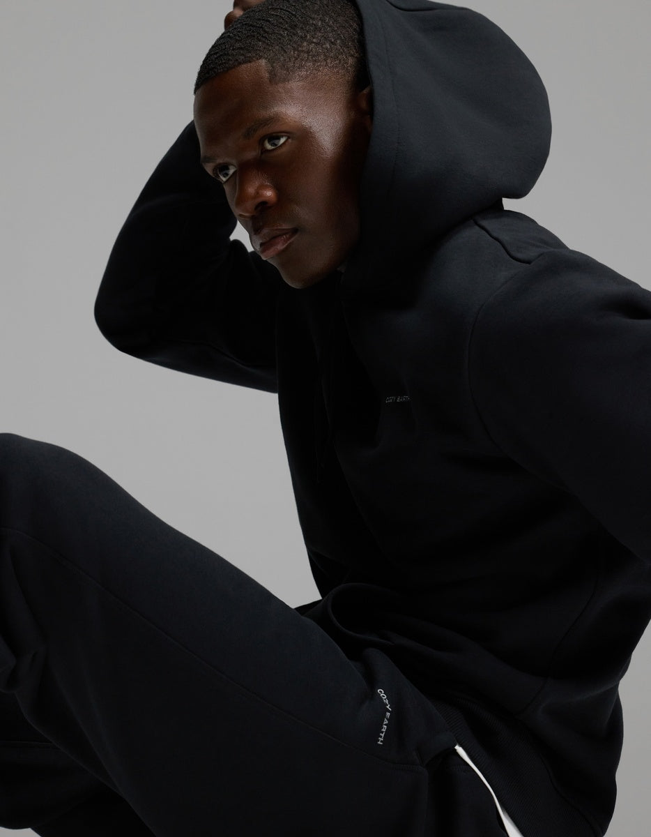 A man wearing Cozy Earth’s Men’s Cityscape Sweatpant in black sits in a relaxed pose against a plain gray background, looking to the side with one hand on his head.|Color:Jet Black