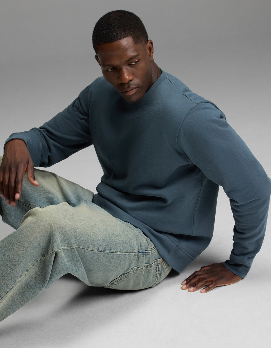 A man models the Cozy Earth Men’s Dylan Crewneck in blue, paired with light-wash jeans. He sits on the floor against a plain, neutral background, looking down with one hand on his knee and the other resting beside him.|Color:Dawn