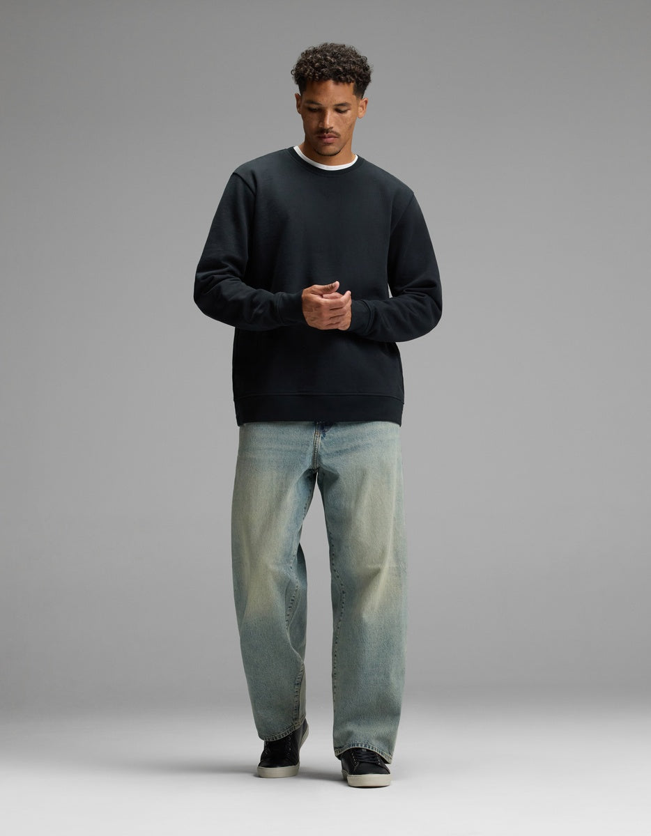 A man stands against a plain gray background, wearing the Cozy Earth Men’s Dylan Crewneck, paired with light blue relaxed-fit jeans and black sneakers. He looks down with his hands clasped together in front of him.|Color:Jet Black