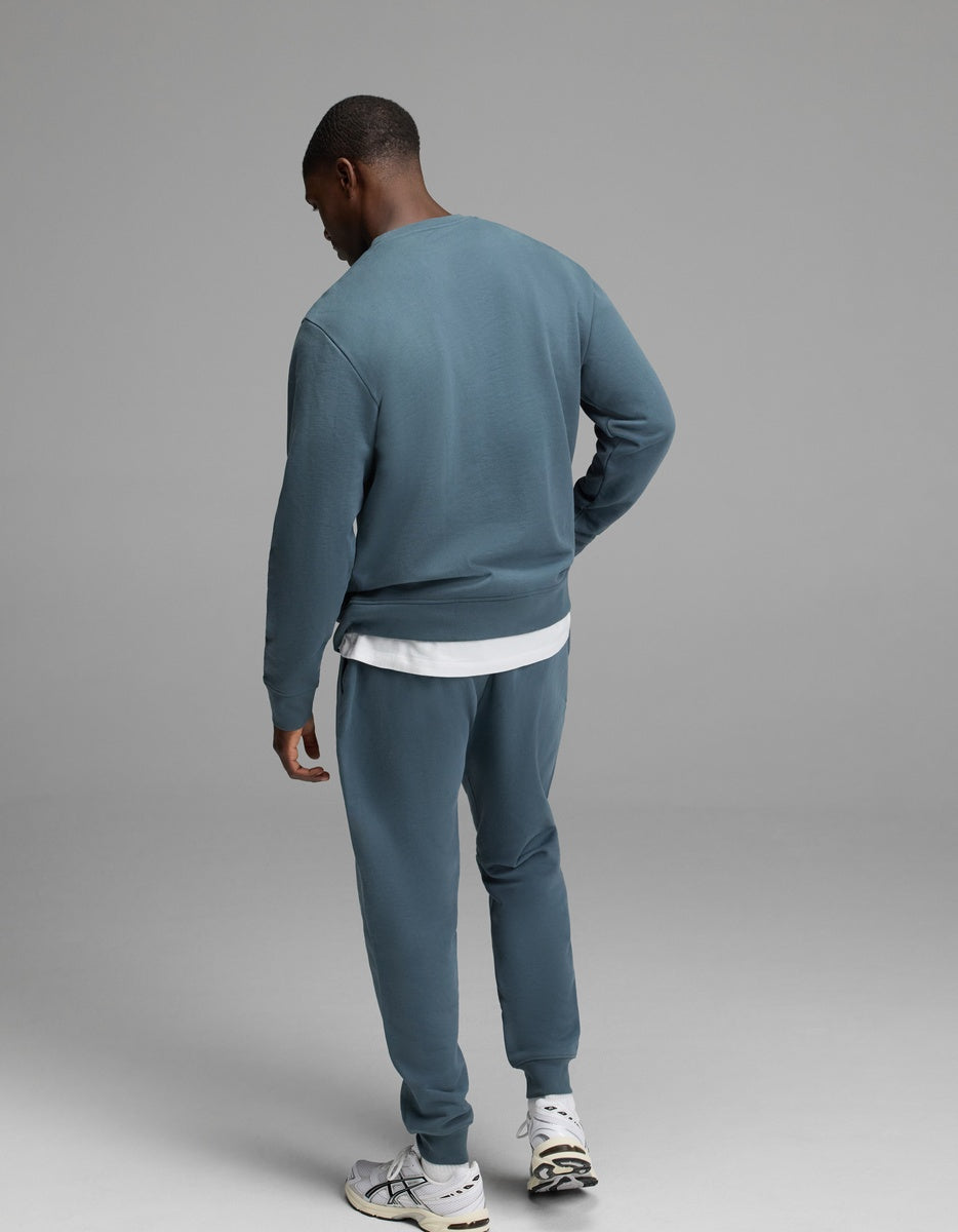 A person stands facing away against a plain gray background, wearing the Cozy Earth Men’s Dylan Crewneck & Jogger Set in blue-green over a white T-shirt and paired with white sneakers.|Color:Dawn