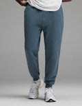 A person is shown from the waist down wearing Cozy Earth’s Men’s Dylan Jogger in blue, paired with a white shirt and white athletic sneakers, standing against a plain gray background.