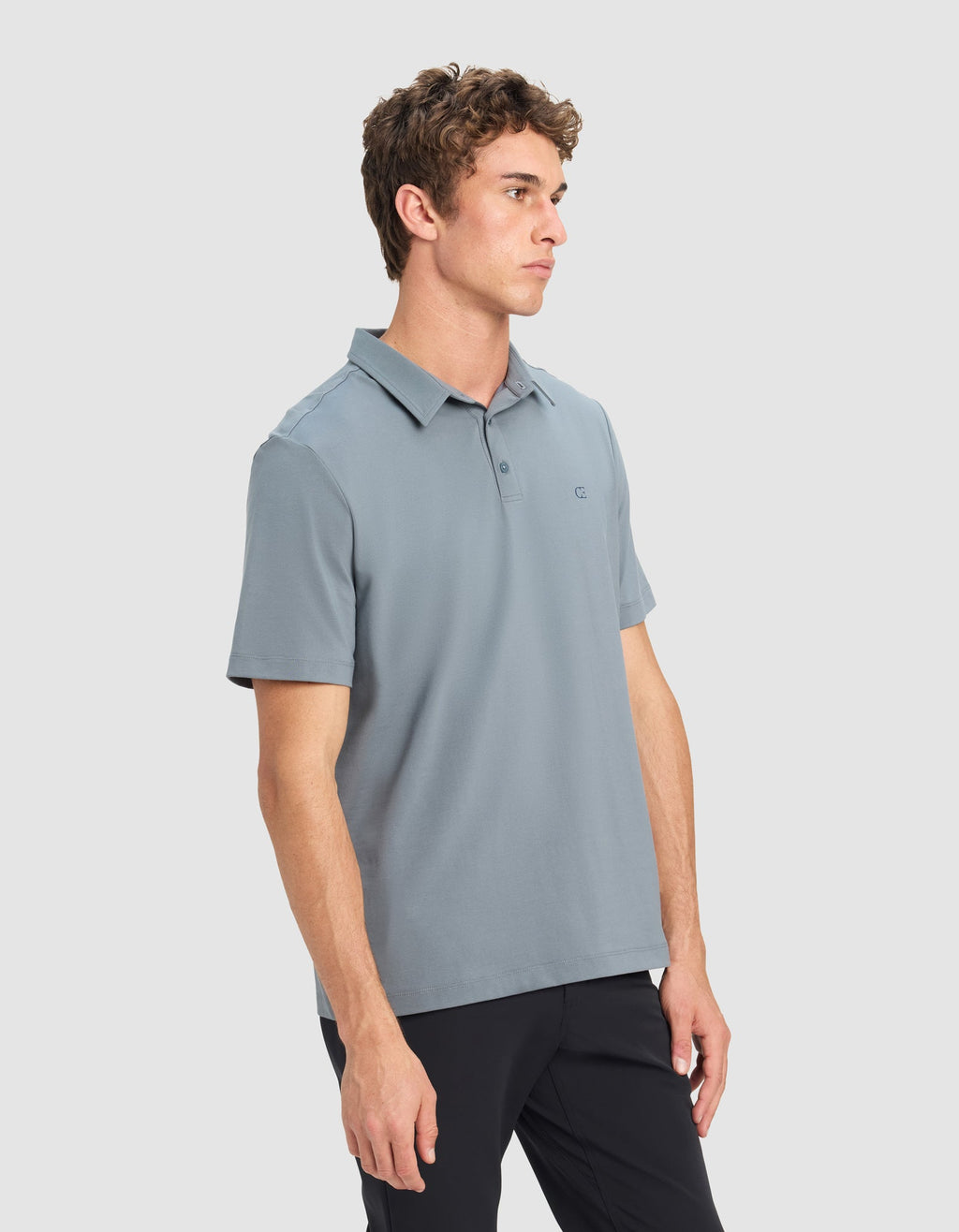 A young man with short curly hair models the Cozy Earth Men's Everyday Polo in light blue and black pants, standing against a plain light gray background and looking off to the side with a neutral expression.|Color:Deep Blue