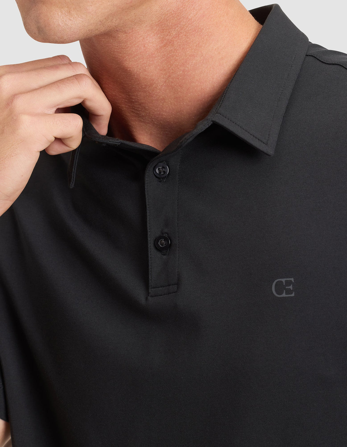 A man wears the Cozy Earth Men's Everyday Polo in black, featuring a two-button collar and a subtle "CE" logo on the chest. His hand is near his neck, and his face is partially visible. |Color:Jet Black
