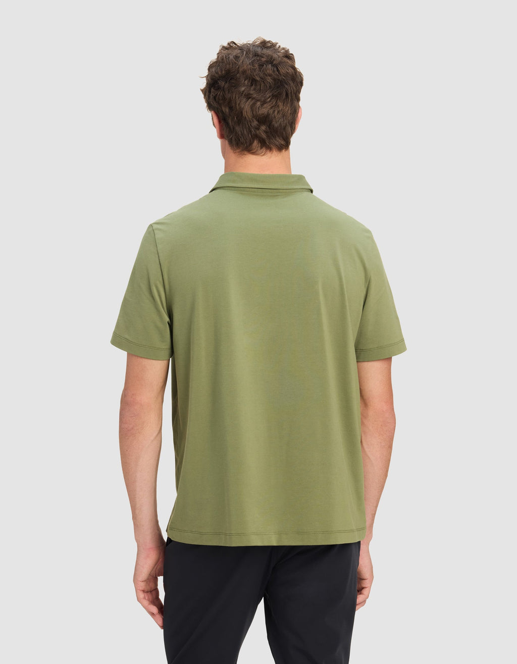 A man is seen from the back, wearing Cozy Earth's Men's Everyday Polo in light olive green and dark pants, set against a plain light gray background.|Color:Juniper
