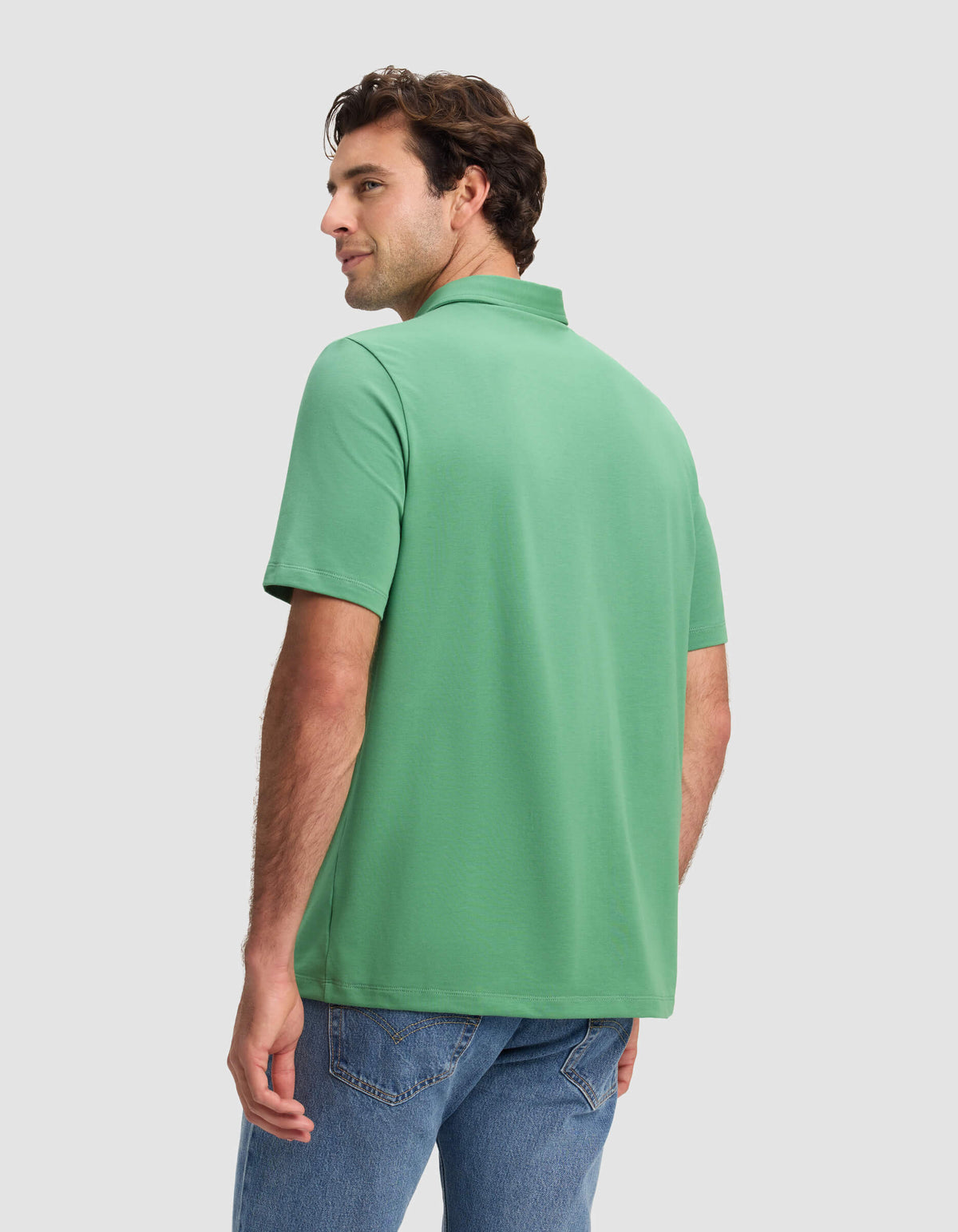 A man stands with his back facing us, turning his head left, wearing the Cozy Earth Men's Everyday Polo (short-sleeve, green) and blue jeans against a plain light gray background.|Color:Vine